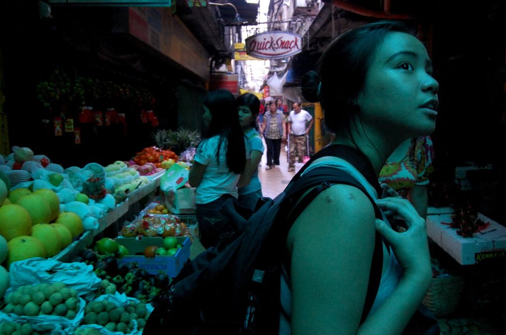 Young Asian woman in a busy street market looking up with curiosity, surrounded by colorful fruit stalls, shoppers, and a neon sign that reads QuickSnack in the dimly lit, narrow alley.