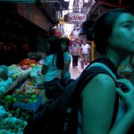 Young Asian woman in a busy street market looking up with curiosity, surrounded by colorful fruit stalls, shoppers, and a neon sign that reads QuickSnack in the dimly lit, narrow alley.